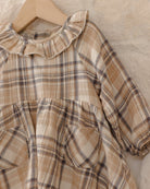 A close-up of a children's plaid dress in beige, brown, and gray tones, hanging on a wooden hanger against a textured beige fabric background.
