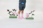 Person wearing pink socks with floral patterns standing between two small plants on a white background