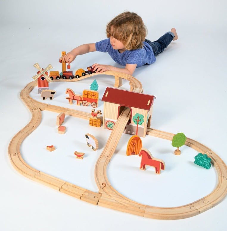 A young child lying on the floor, reaching out to play with a wooden farmyard-themed train set complete with tracks, a red barn, a windmill, and various animal figures.