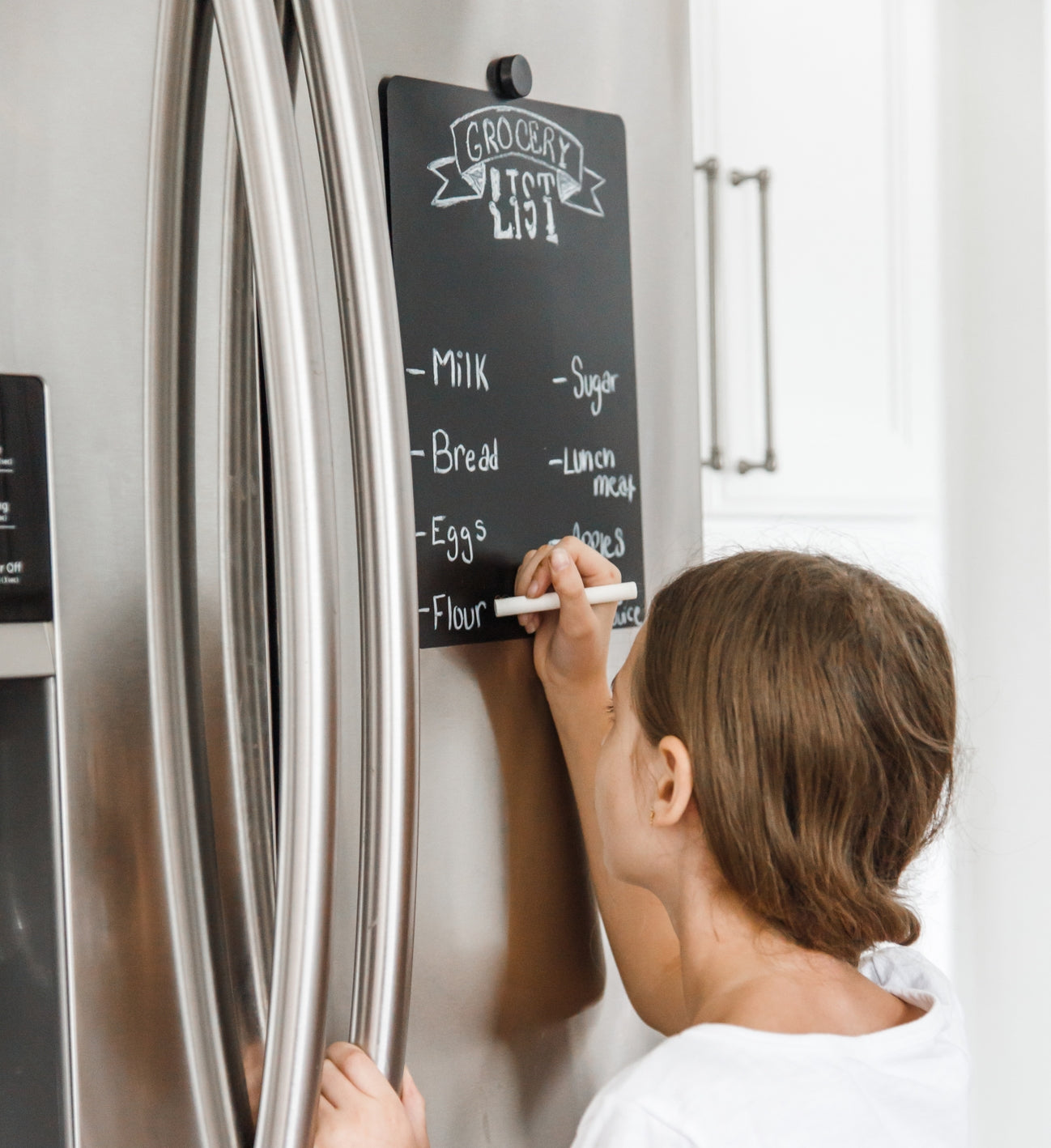 Child writing on a chalkboard attached to a refrigerator with a 'Grocery List' written on it.