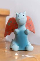 A small blue dragon figurine with orange wings decorated with blue stars, sitting on a wooden surface with warm fairy lights in the foreground.