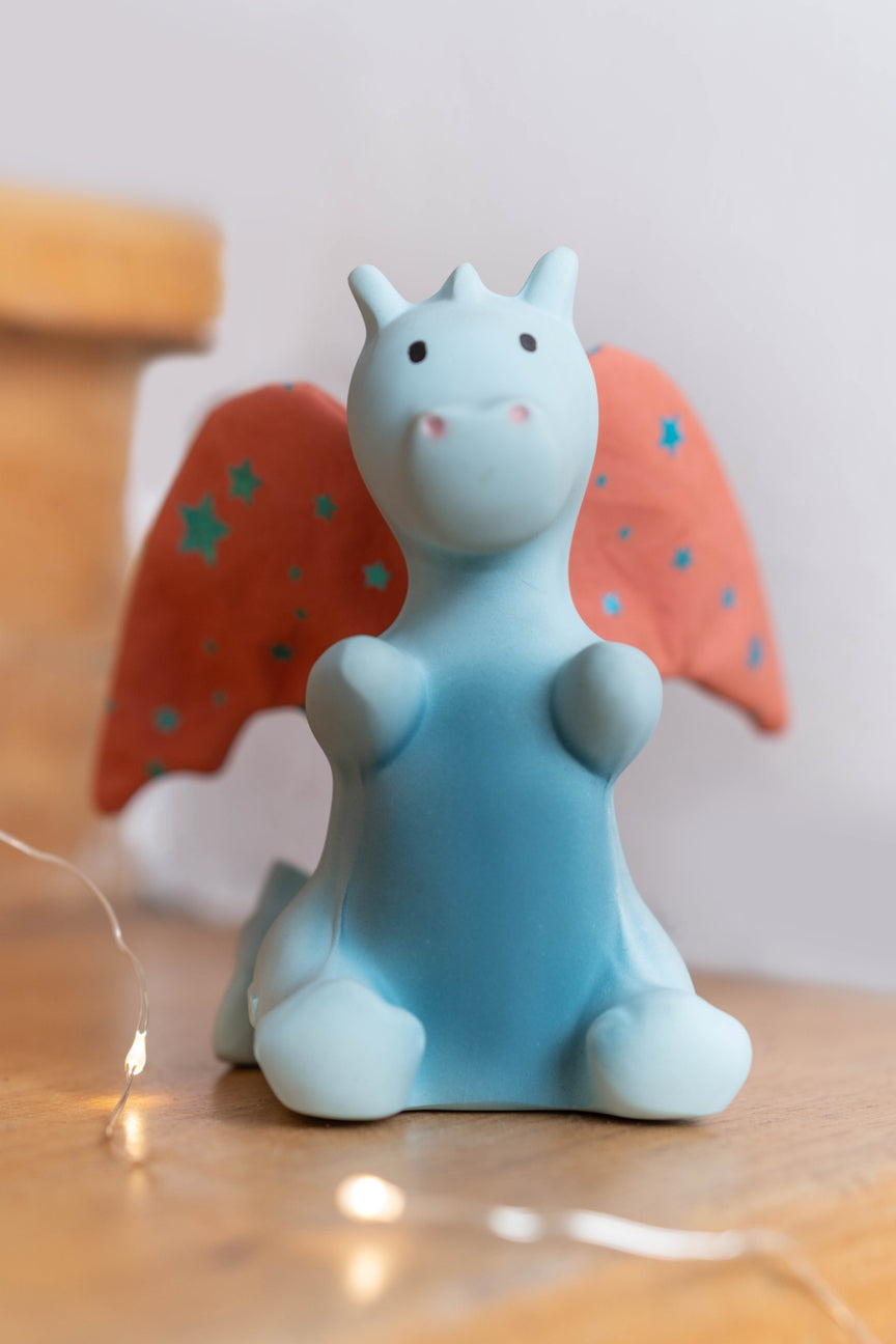 A small blue dragon figurine with orange wings decorated with blue stars, sitting on a wooden surface with warm fairy lights in the foreground.