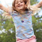 Child wearing a colorful rainbow-patterned shirt with a logo, standing outdoors with arms outstretched.