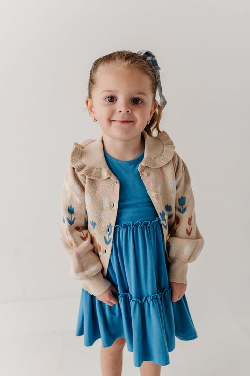 Young girl wearing a beige jacket with floral patterns and a blue dress on a plain background