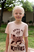 A young girl wearing a light peach-pink graphic t-shirt with a brown cartoon kitten and the text 'BRING ME All the KITTIES'.