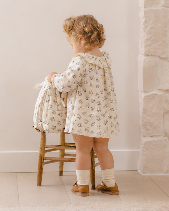 A toddler stands on a wooden stool wearing a cream floral dress and holding a matching floral fabric bag, photographed in a softly lit indoor room with a light wall and stone column.