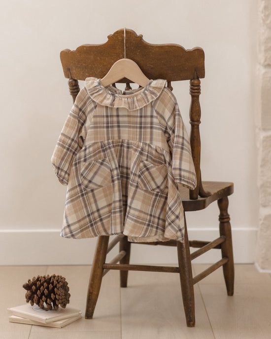 A toddler girl's long-sleeve plaid dress in beige, brown, and white hangs on a wooden hanger from a rustic wooden chair, with a pinecone and books on the floor nearby.