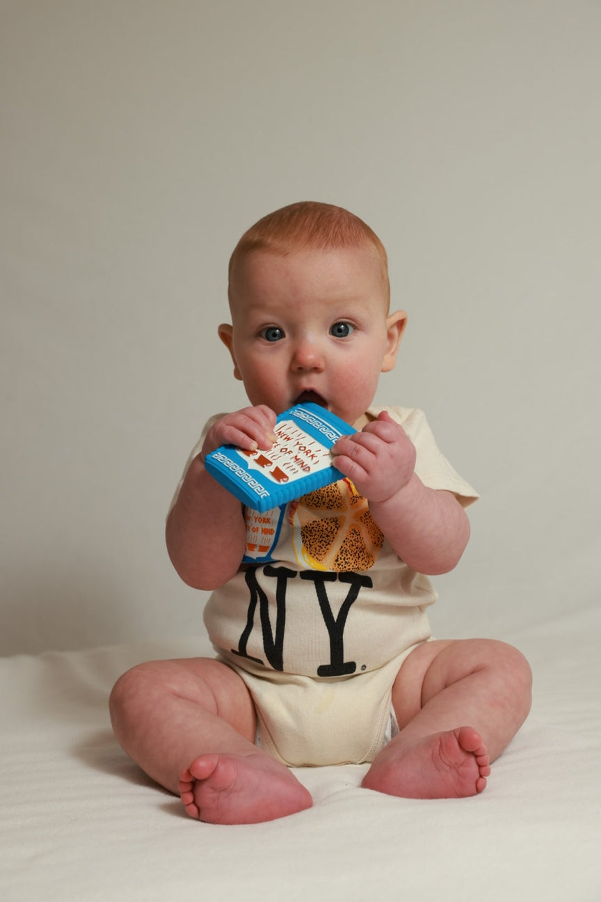 A baby sitting on a white surface against a light beige background, wearing a cream-colored onesie with 'NY' printed in black and holding a blue teething toy.