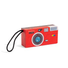 A red camera-shaped speaker by moulinsroty, featuring a black wrist strap, a blue lens graphic, and a retro rainbow stripe on the side, shown on a white background.