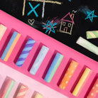 A top-down view of pastel chalk sticks arranged on a bright pink surface, with a black chalkboard in the background featuring colorful chalk drawings.