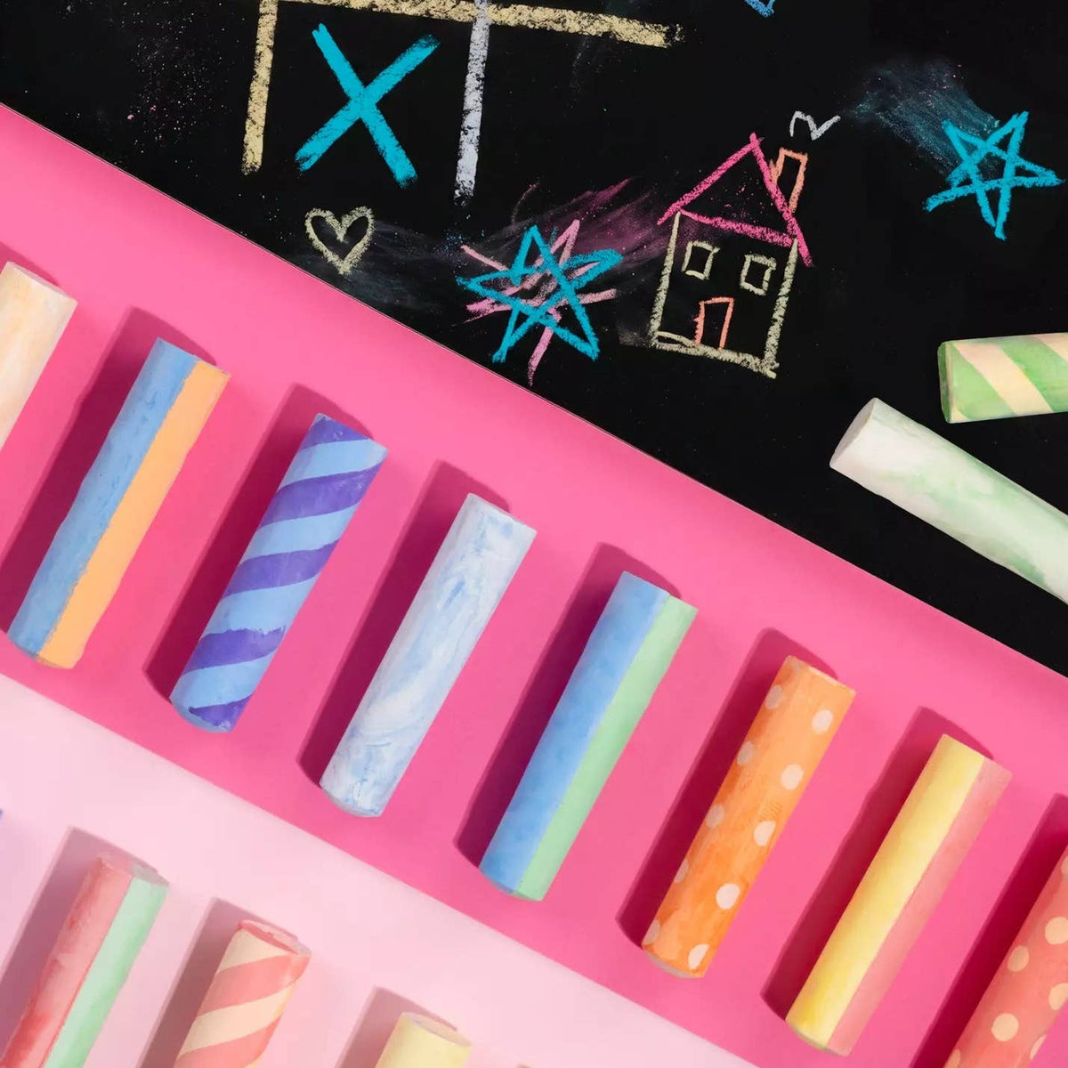 A top-down view of pastel chalk sticks arranged on a bright pink surface, with a black chalkboard in the background featuring colorful chalk drawings.