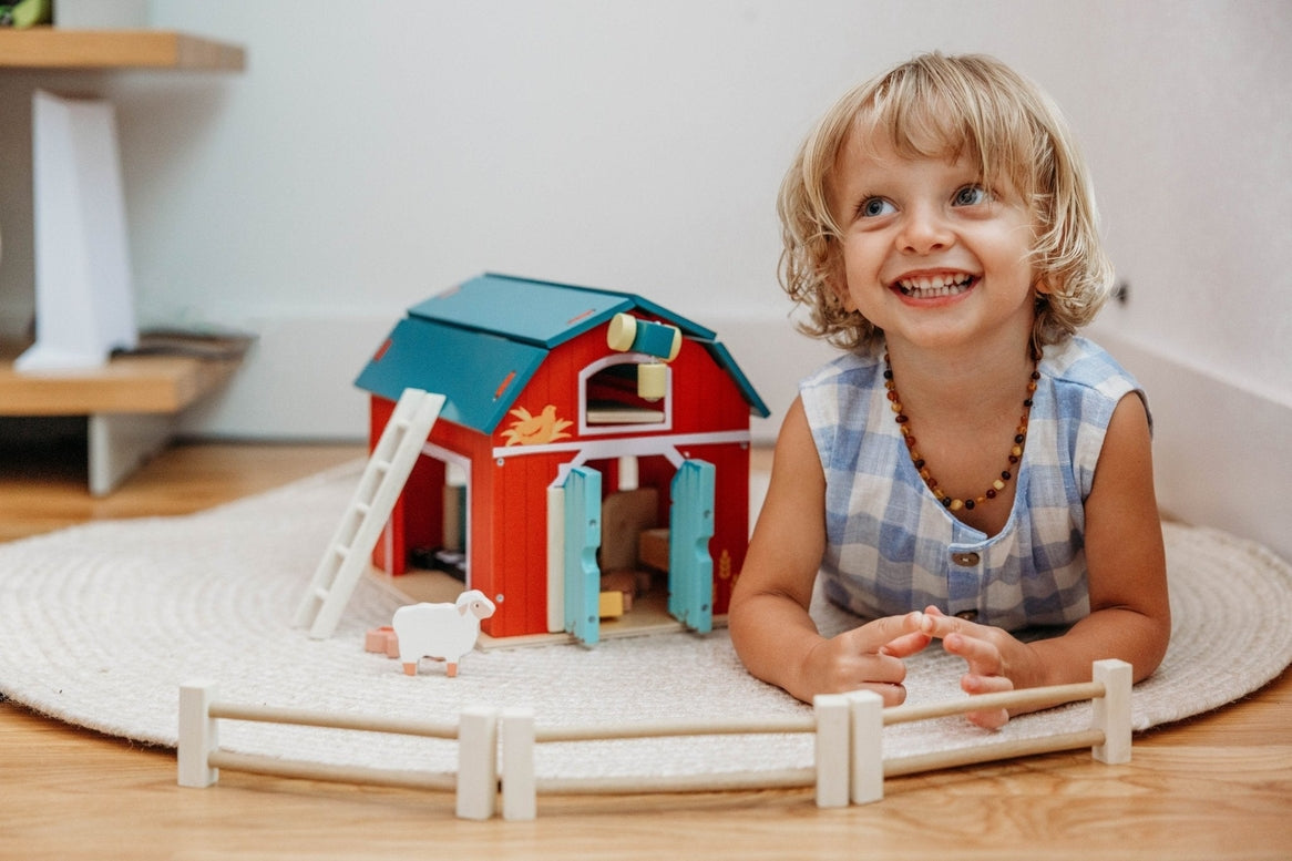 A young blonde-haired child, wearing a blue and white gingham sleeveless collared shirt, smiles happily while playing on the floor with the Wooden Big Red Barn Playset by Mentari, surrounded by the barn, fences, and small wooden animal figures.