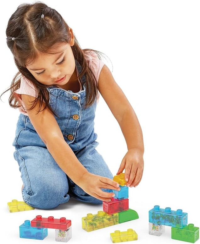 A young girl wearing denim overalls kneeling on a white background and stacking colorful, translucent Jelly Blox sensory blocks.