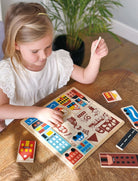 A young girl with blonde hair tied back sits at a wooden table, smiling as she plays with the Mentari Wooden Town Puzzle, holding a puzzle piece in her hand while assembling the colorful town scene.