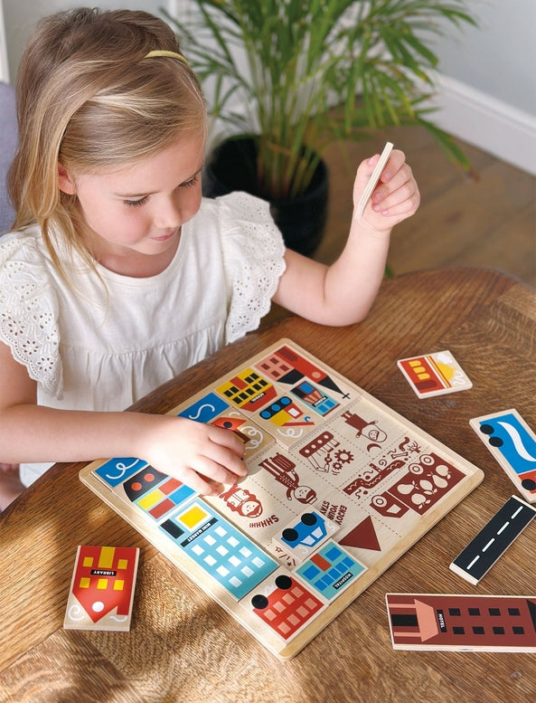 A young girl with blonde hair tied back sits at a wooden table, smiling as she plays with the Mentari Wooden Town Puzzle, holding a puzzle piece in her hand while assembling the colorful town scene.