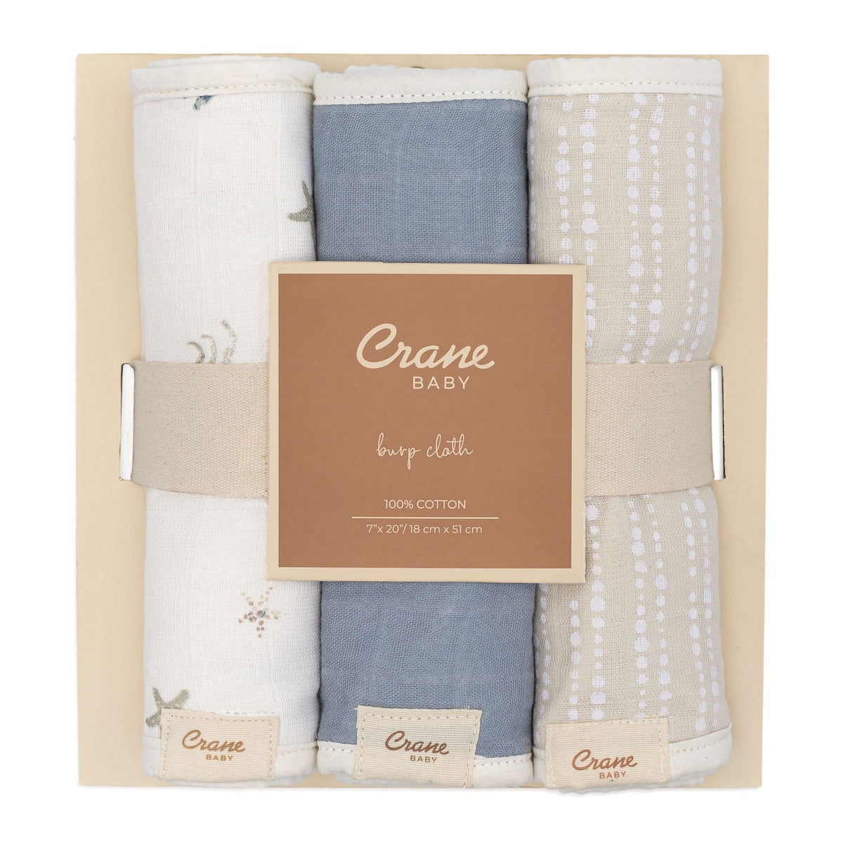 A boxed set of three baby burp cloths from Crane Baby, neatly folded and presented in a light beige gift box with a central label. The cloths are in soft neutral tones: one off-white with delicate gray botanical prints, one dusty blue, and one light beige with white dotted patterns.