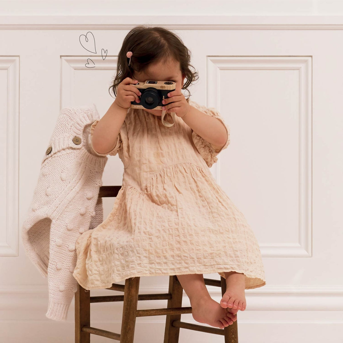 Child in a beige dress holding a camera against a white wall.