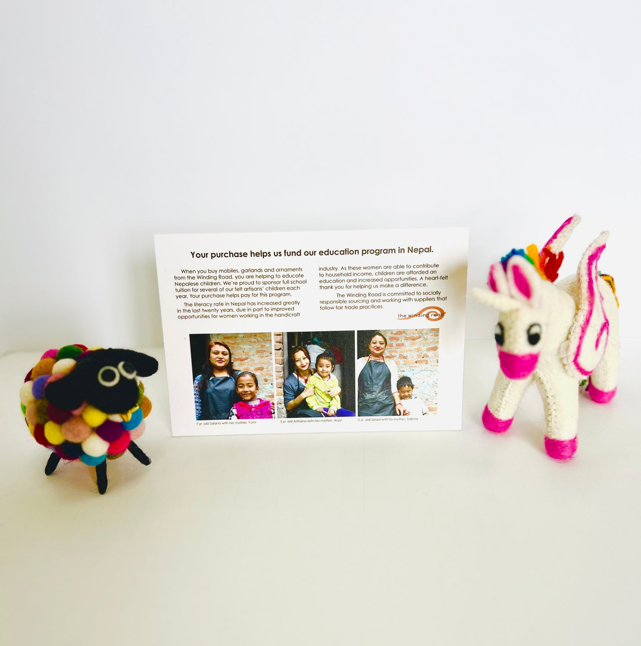 A handmade felted wool toy set displayed on a white surface against a white background, featuring a colorful fluffy sheep, a white unicorn with rainbow mane and tail, and a printed informational card in the center.