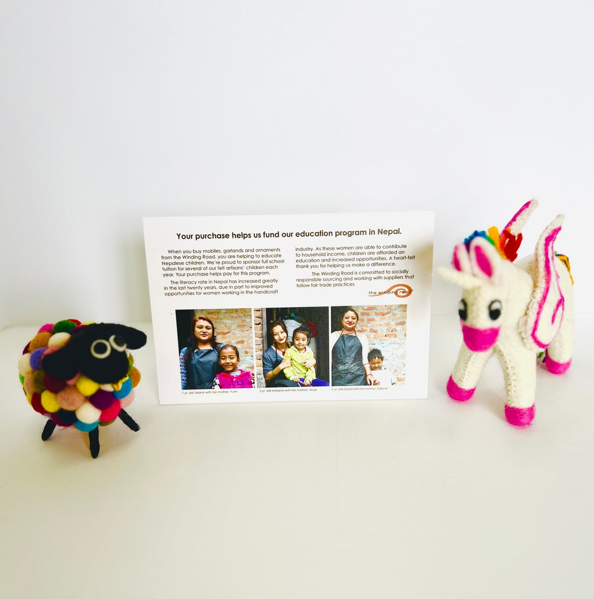 A handmade felted wool toy set displayed on a white surface against a white background, featuring a colorful fluffy sheep, a white unicorn with rainbow mane and tail, and a printed informational card in the center.