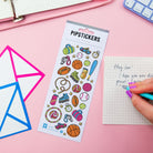A sheet of colorful pip stickers labeled 'PIPPSTICKERS' is displayed on a pink desk. The sticker sheet contains various sports-themed illustrations like a basketball, baseball, bat, and sneakers.