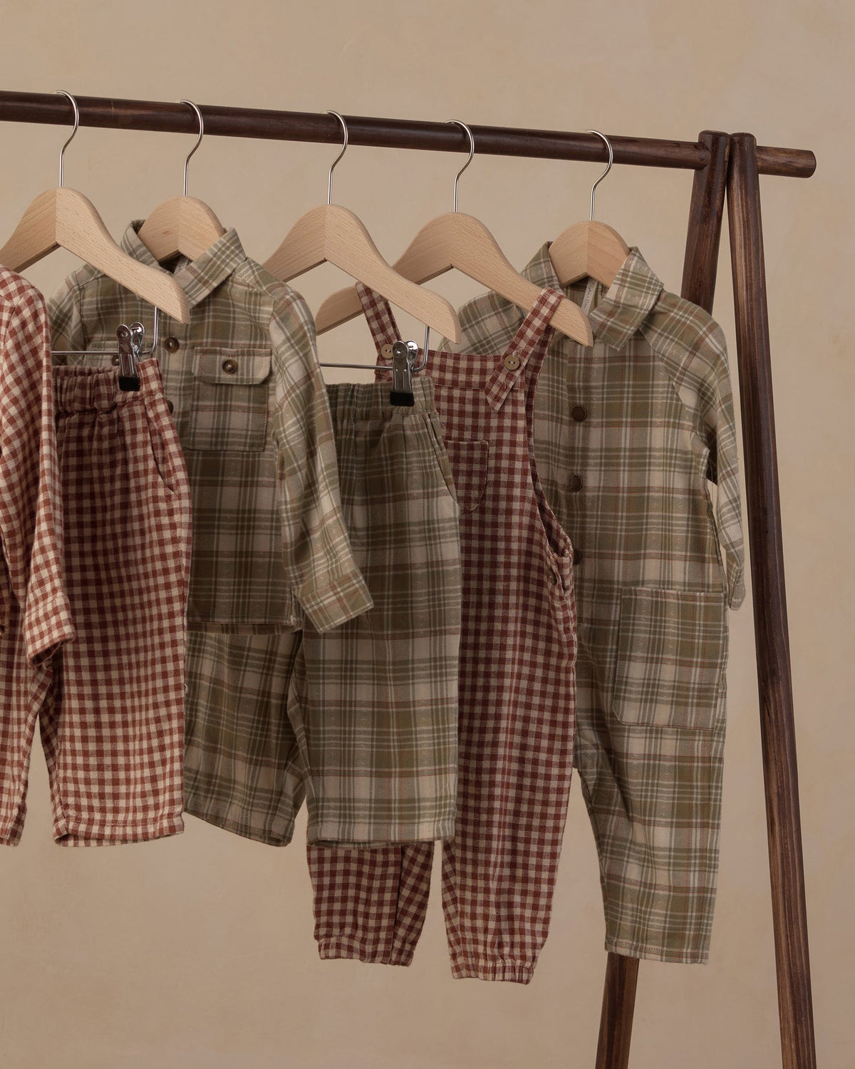 Three children's two-piece clothing sets hanging on wooden hangers from a dark wood rack. The sets include plaid button-up shirts and matching overalls in green/olive and brown/red checks, set against a plain beige background.