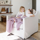 Two young girls sitting on a white armchair with a purple blanket in a cozy living room.