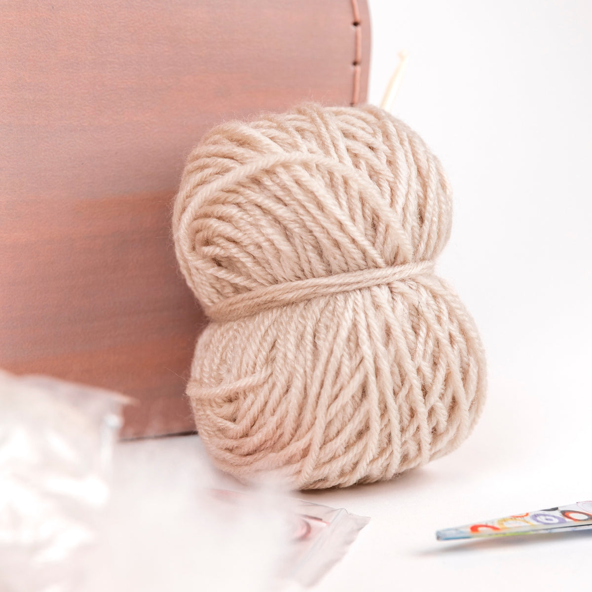 Two skeins of beige yarn on a white surface with a pink cushion in the background.