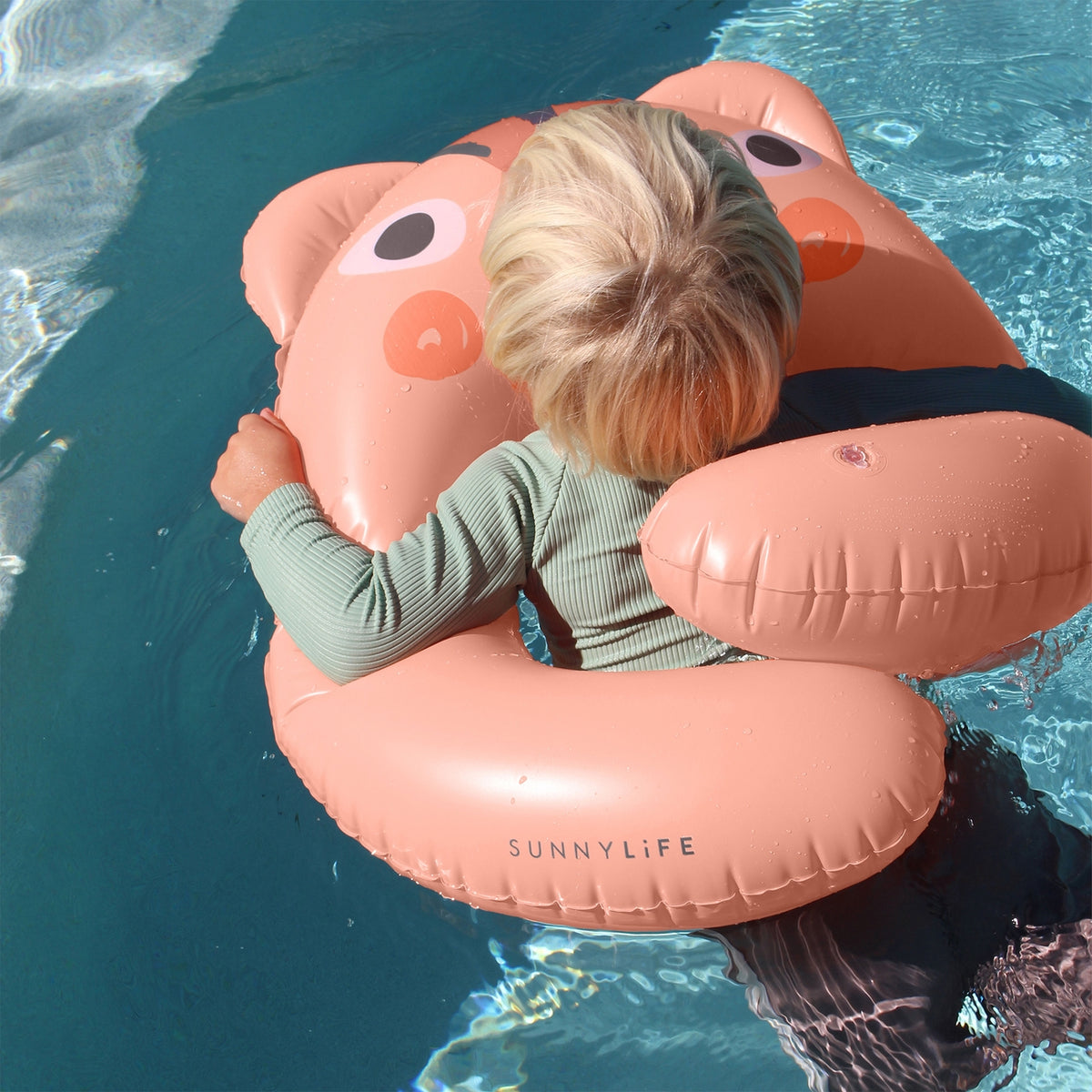 Child on a pink inflatable animal float in a pool with 'SUNNYLIFE' branding.