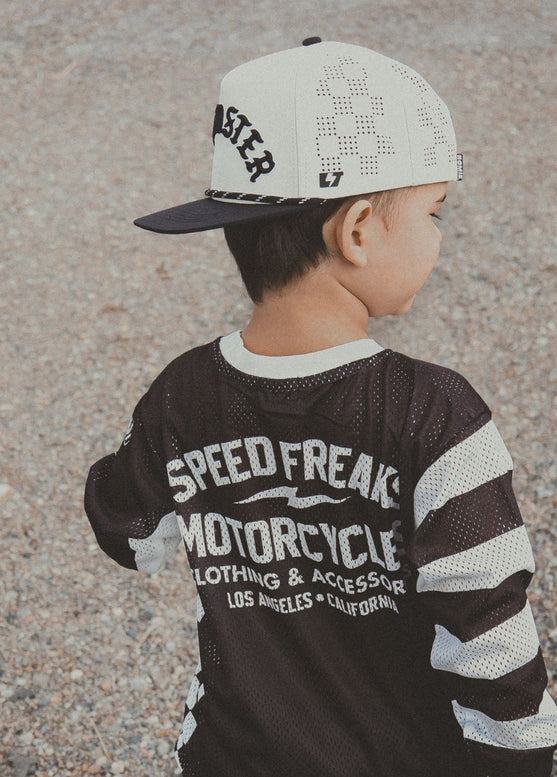 A child wearing a black and white mesh long-sleeve shirt with 'SPEED FREAK MOTORCYCLE' printed on the back, paired with a white and black baseball cap, photographed outdoors on a gravel surface.