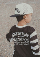 A child wearing a black and white mesh long-sleeve shirt with 'SPEED FREAK MOTORCYCLE' printed on the back, paired with a white and black baseball cap, photographed outdoors on a gravel surface.
