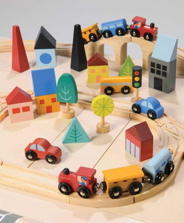 A top-down, angled view of a wooden toy train set on a light surface, featuring a curved wooden track layout with a blue train engine and yellow cargo cars. Colorful wooden town buildings, including a red house, blue structures, and abstract trees, surround the tracks alongside a yellow traffic light, a red toy car, and a yellow toy truck.