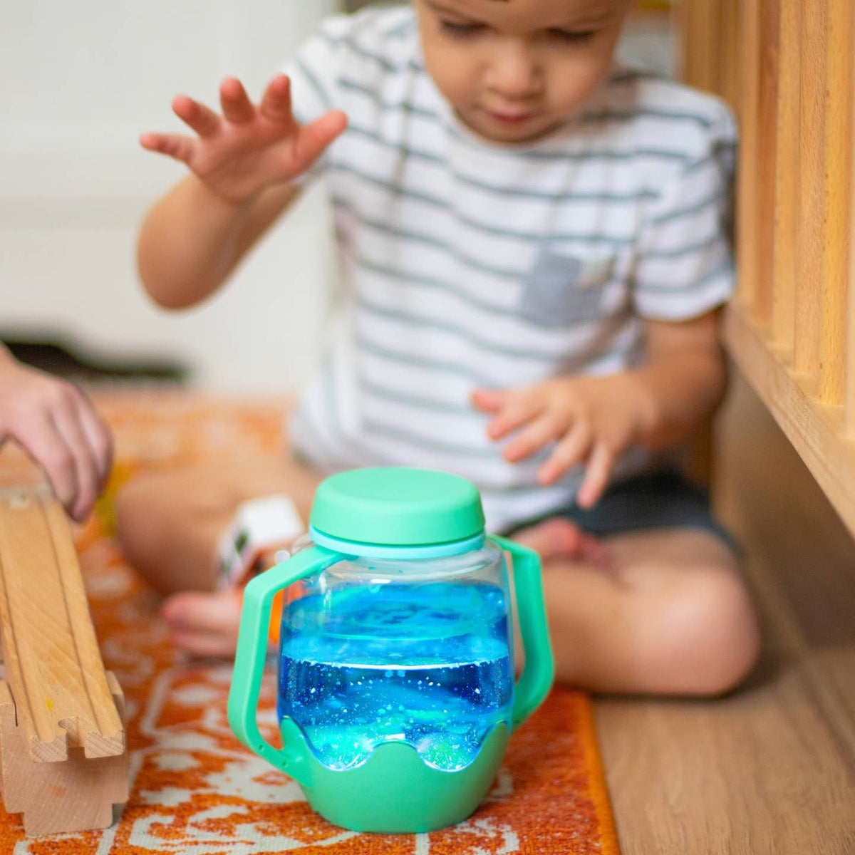 Child playing with a green and clear container filled with blue liquid on a wooden floor.