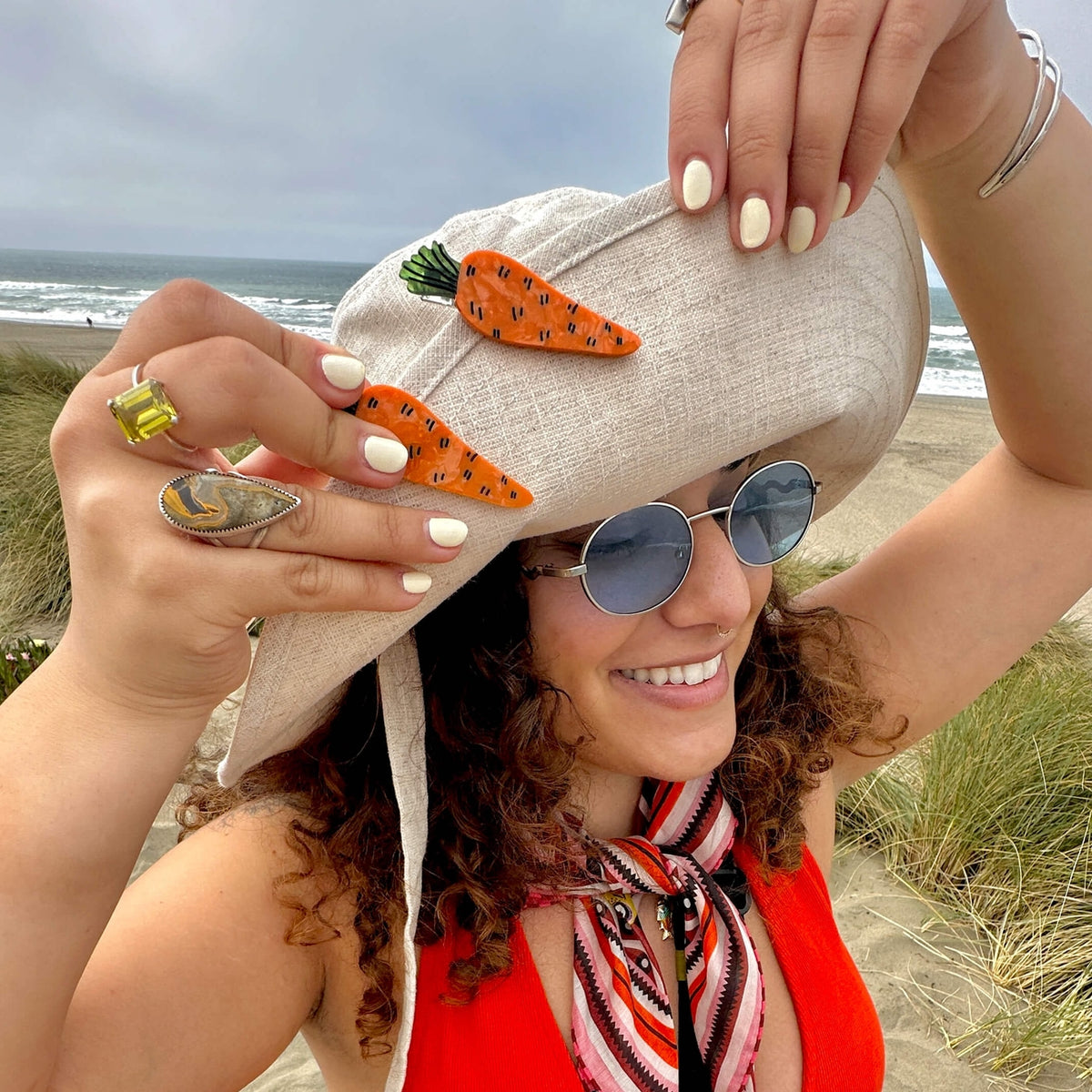 Woman on a beach wearing a hat with carrot decorations, surrounded by ocean and grass.