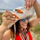Woman on a beach wearing a hat with carrot decorations, surrounded by ocean and grass.
