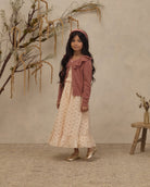 A young girl stands in a styled studio setting wearing a long cream dress with a small floral print, a dusty rose cardigan, and a matching headband. The background features a textured beige wall, dried botanical branches, and a small wooden stool.