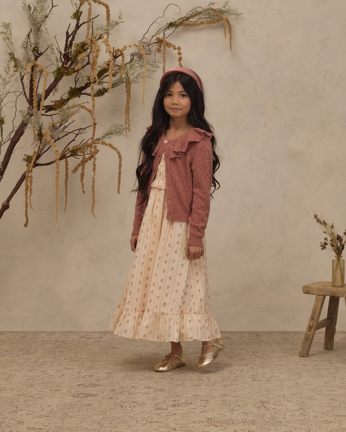 A young girl stands in a styled studio setting wearing a long cream dress with a small floral print, a dusty rose cardigan, and a matching headband. The background features a textured beige wall, dried botanical branches, and a small wooden stool.