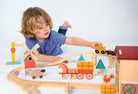 A young child playing on the floor with a wooden farmyard-themed train set, placing a small wooden train onto the tracks next to a red barn and windmill.