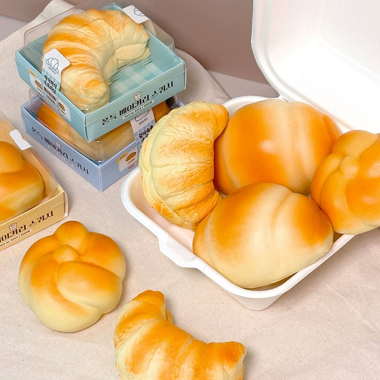 A styled product display of various bakery items, including golden-brown croissants, round buns, and braided bread, with some packaged in boxes and others arranged on a beige fabric surface.