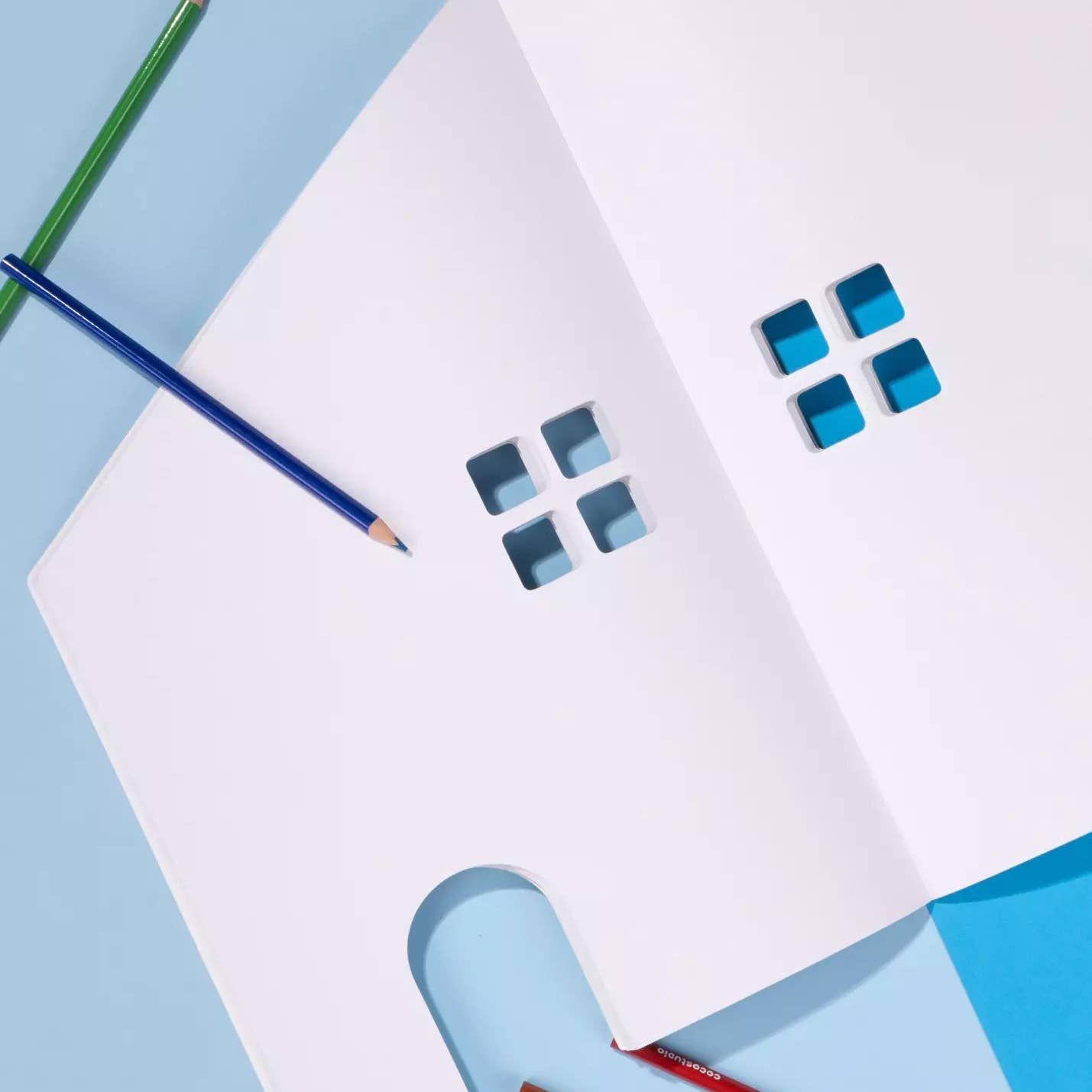 A white house-shaped paper craft or card with cutout window and door shapes, arranged on a light blue background with colored pencils.