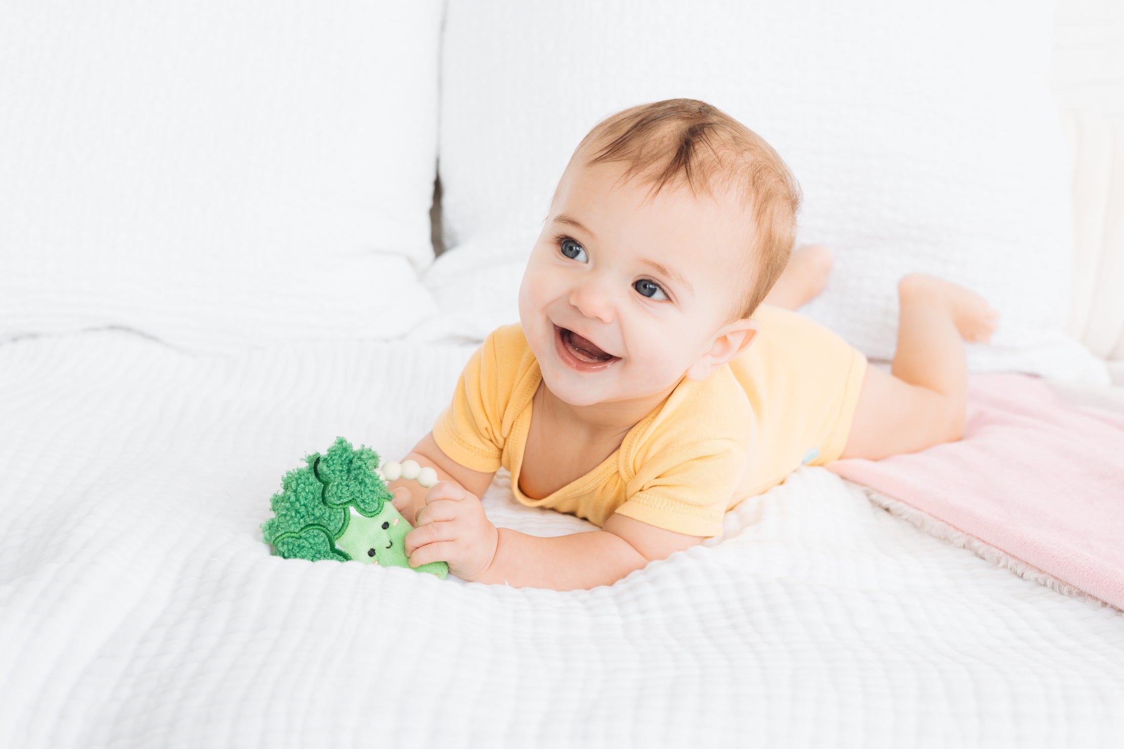 Baby lying on a white blanket with a green toy, smiling.