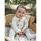 A baby sitting in a woven rattan bassinet outdoors, wearing a cream-colored footed zip-up sleeper with owl and bird prints.
