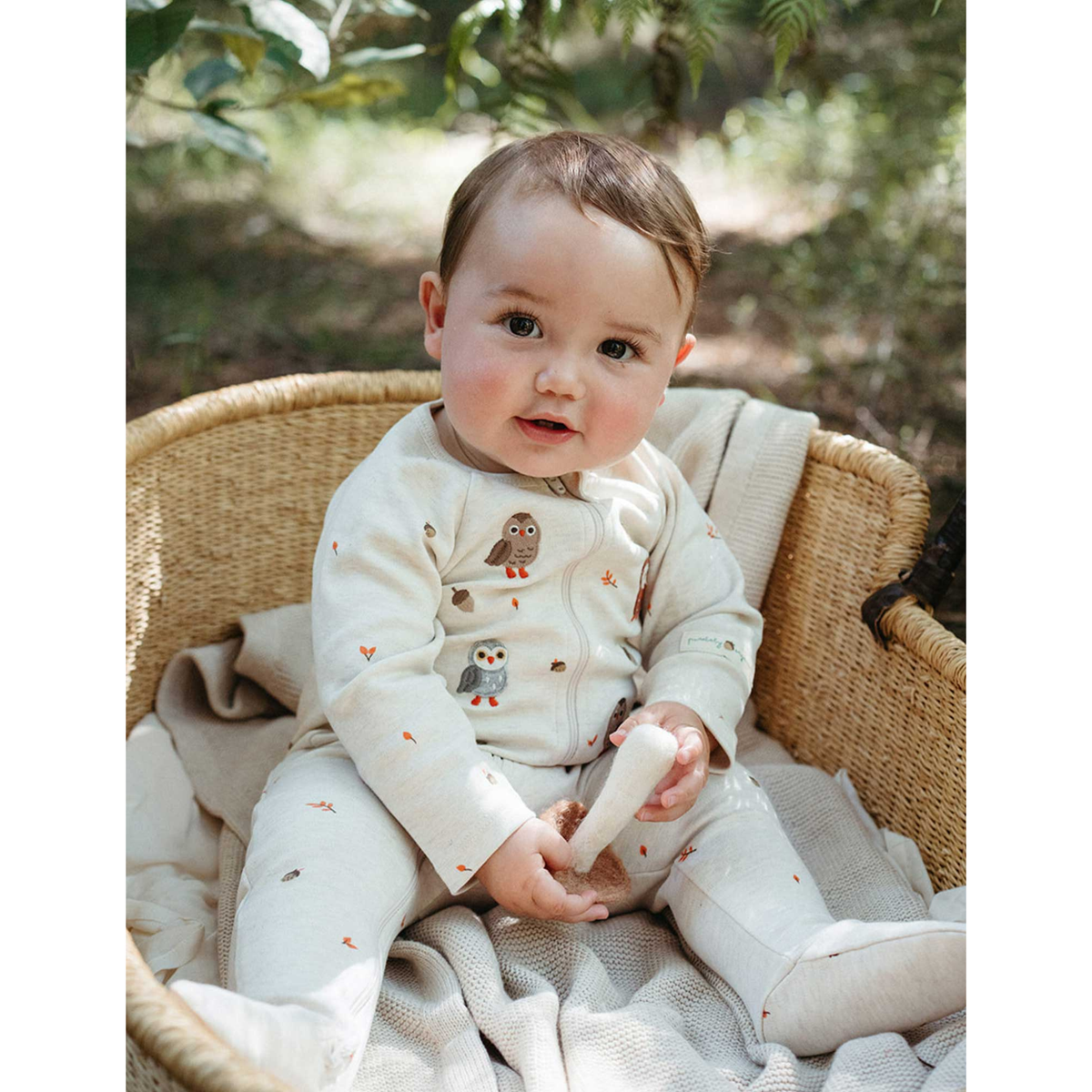 A baby sitting in a woven rattan bassinet outdoors, wearing a cream-colored footed zip-up sleeper with owl and bird prints.