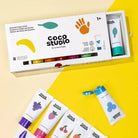 Coco Studio Scented Finger Point set in a white box with a sliding drawer, shown on a split cream and bright yellow background with several white tubes featuring colorful fruit illustrations.
