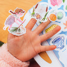 Hand holding a colorful sticker with a fairy design on a book page.