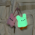 Two small plush bunny keychains hanging from a beige backpack. One bunny is pink and the other is glowing mint green with a small orange shape peeking out from behind, both with simple black facial expressions.