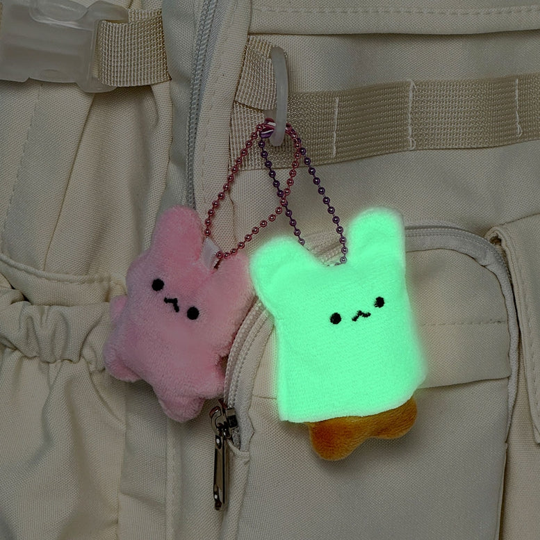 Two small plush bunny keychains hanging from a beige backpack. One bunny is pink and the other is glowing mint green with a small orange shape peeking out from behind, both with simple black facial expressions.