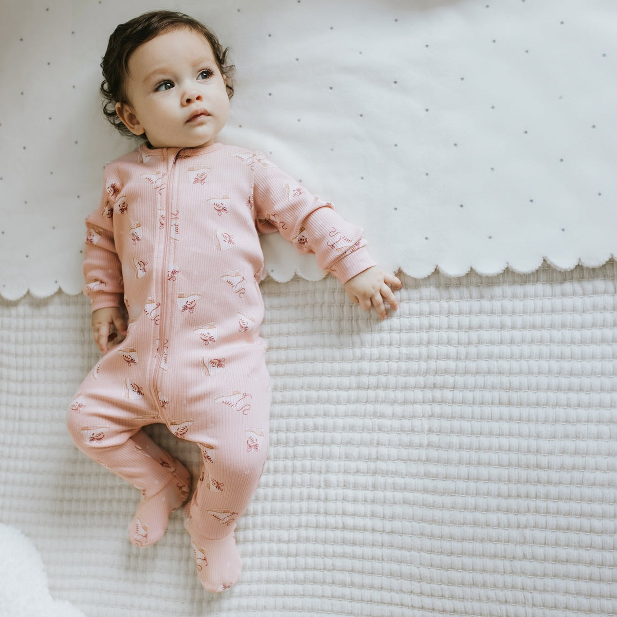 A baby lying on a textured white blanket wearing a pink footed zip-up sleeper with a small floral print.