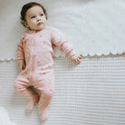 A baby lying on a textured white blanket wearing a pink footed zip-up sleeper with a small floral print.