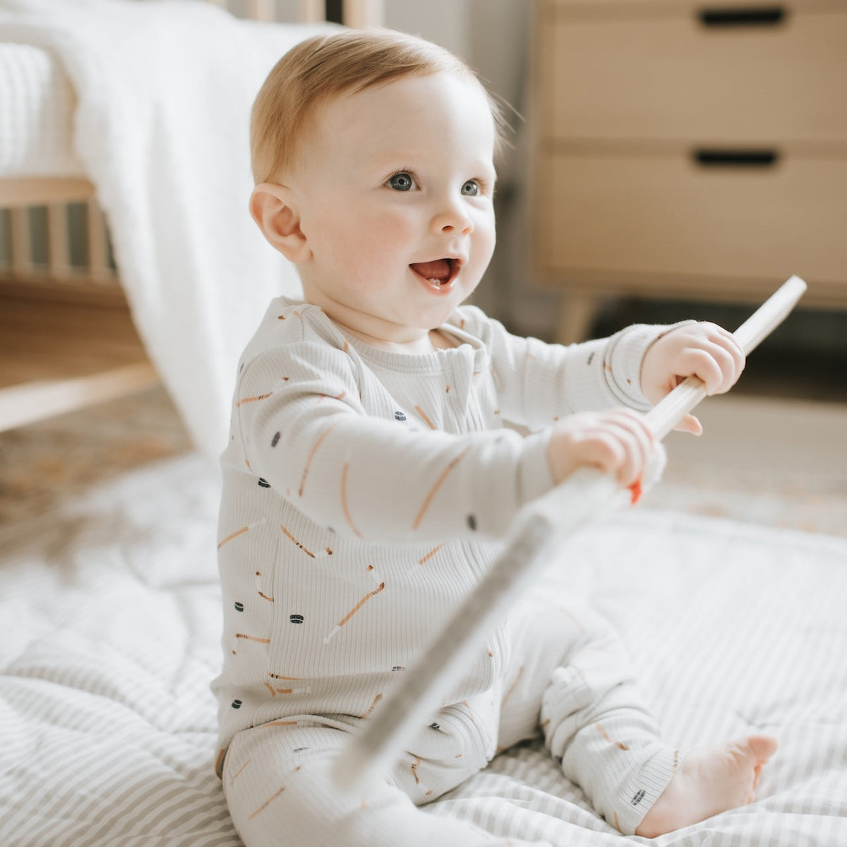 A baby sitting on a white blanket in a nursery, wearing a cream ribbed long-sleeve footless onesie with a small pencil print and holding a white toy.