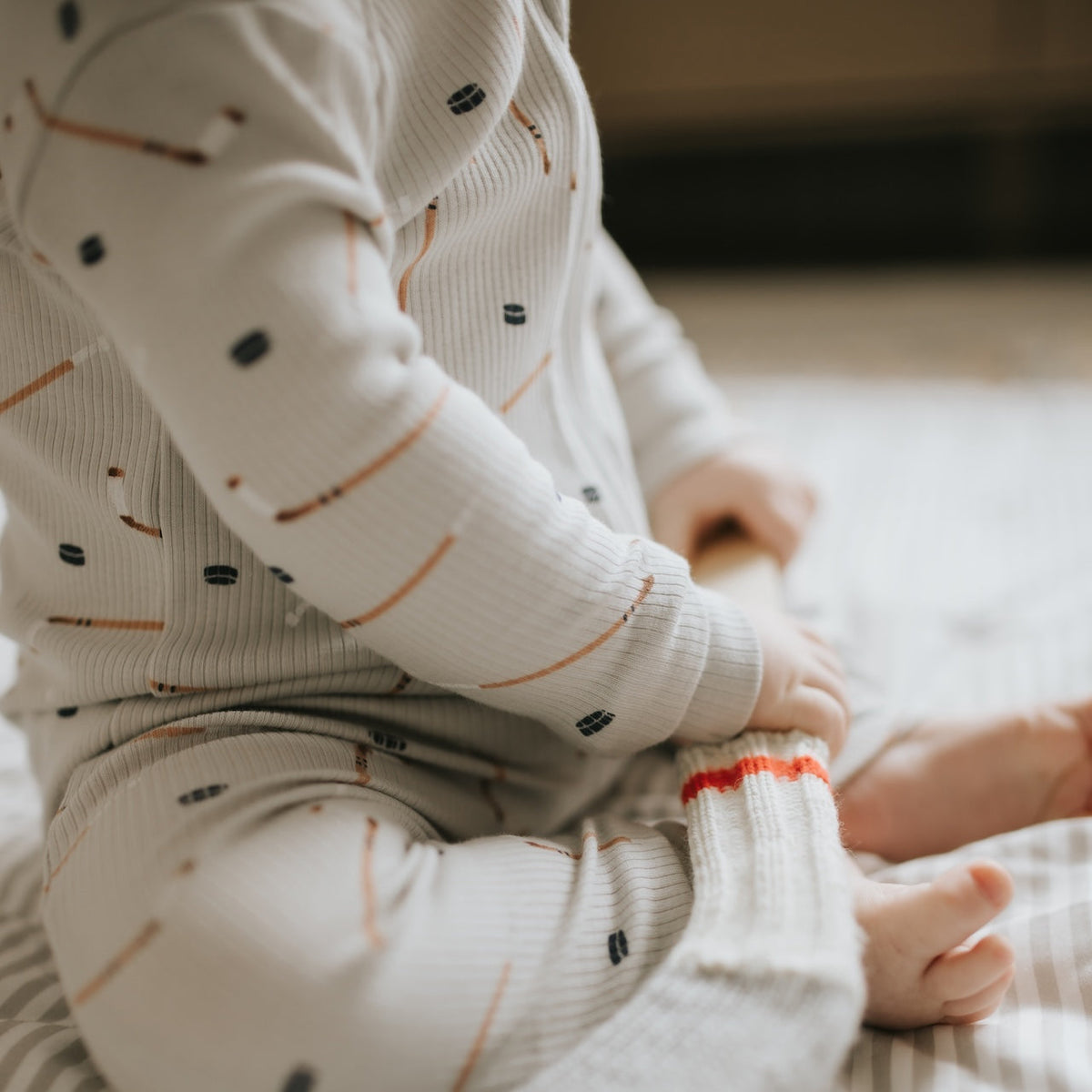 A baby sitting on a white blanket in a nursery, wearing a cream ribbed long-sleeve footless onesie with a small pencil print and holding a white toy.